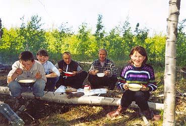 Привал, все сидят в тени возле ствола молодой берёзы и обедают. У всех в руках тарелки и веселые лица.
