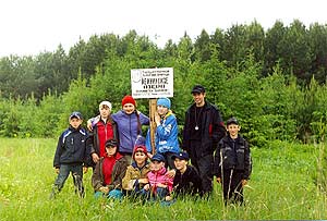 Туристическая группа школы возле таблички перед Лежнинским озером в Пижанском районе. Табличка стоит посреди зеленого луга, по бокам и в переди стоят и сидят походчики. Сзади молодые елки. Светло-серое небо.